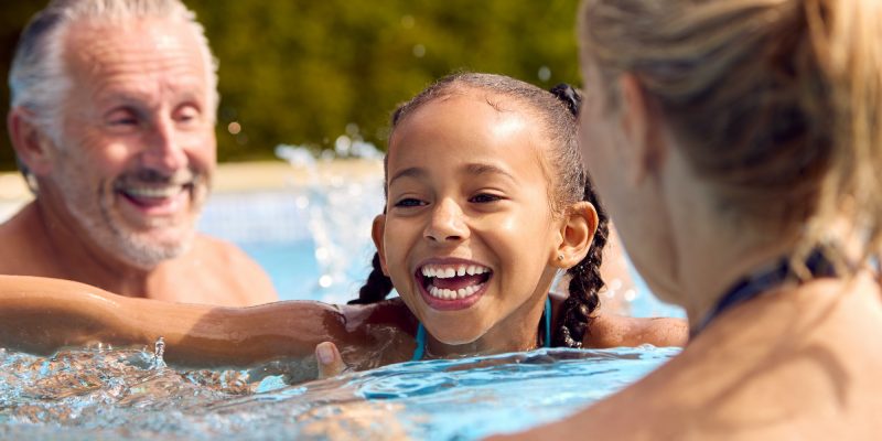 Grandparents Teaching Granddaughter To Swim On Family Summer Holiday In Pool